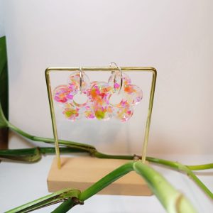 Par de pendientes de resina epoxi transparentes con forma de flor. Adornados con trozos de resina en tonos vibrantes como el rosa, naranja y verde neón. Mostrados en un soporte dorado.